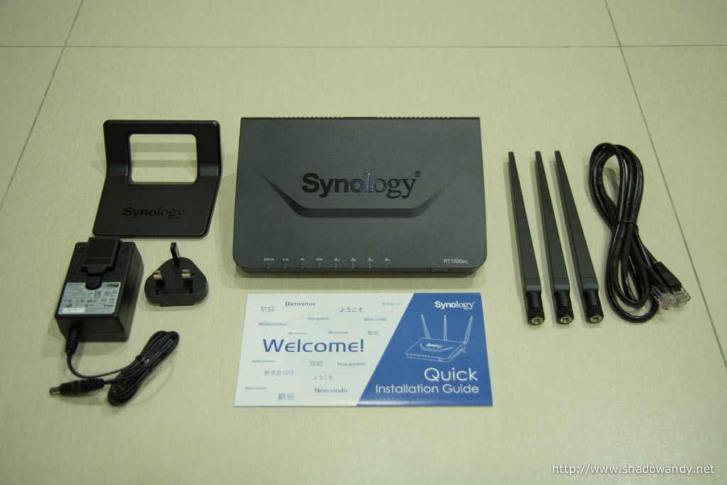 The Synology RT1900ac laid out: RT1900ac WiFi router, stand attachment, power adaptor, dual-band antennas, ethernet patch cord and quick installation guide.